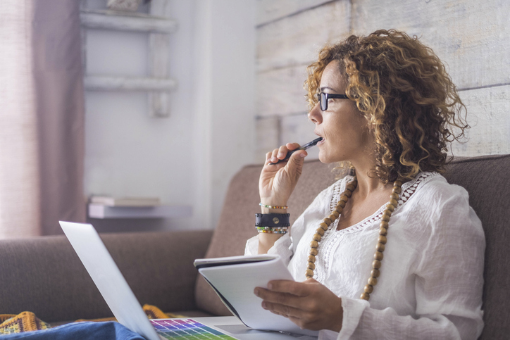 Woman on couch thinking with laptop, notebook and pen in hand.